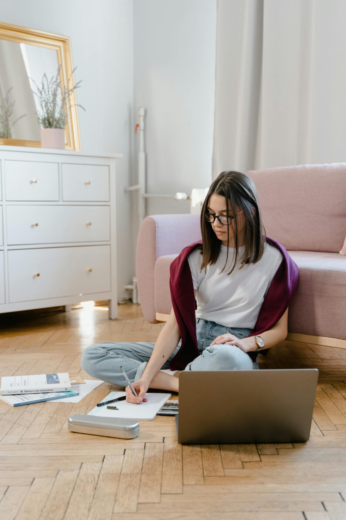 A teenage girl sitting on the floor with a laptop, writing in her notebook